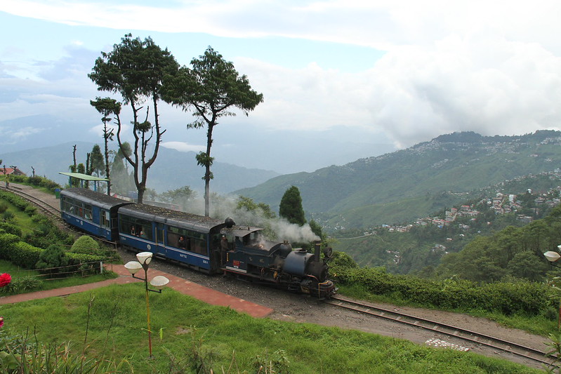 Darjeeling toy train moving along mountain tracks with green hills and misty valleys in the background.