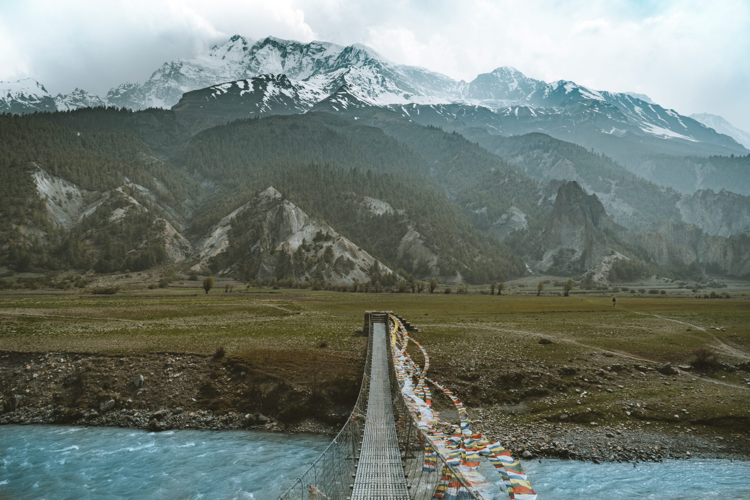 Wooden suspension bridge over blue river with prayer flags and snow-covered Himalayan mountains in background.