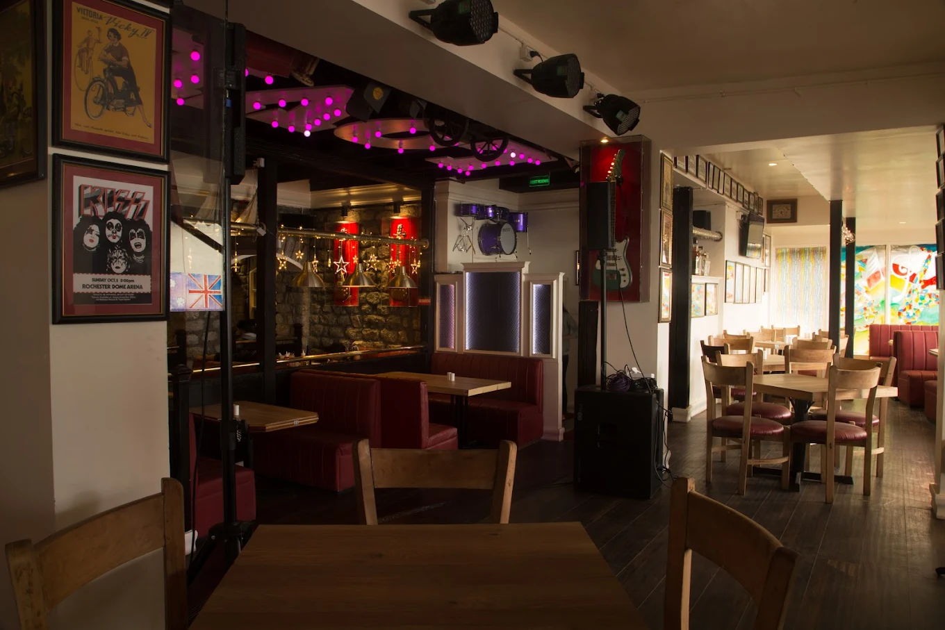 Warmly lit café interior with wooden tables, red booths, framed music posters, and stage lighting.